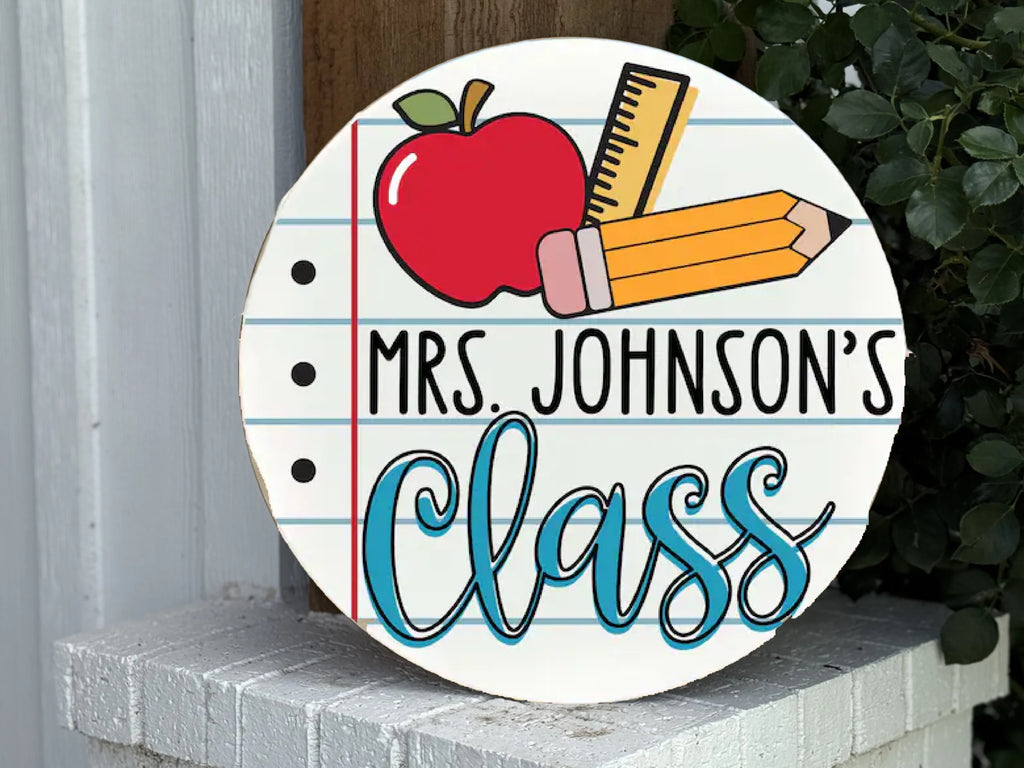 A round sign designed like notebook paper reads Mrs. Johnsons Class with illustrations of an apple, ruler, pencil, and eraser above the text. The sign is propped on a white brick surface near greenery.
