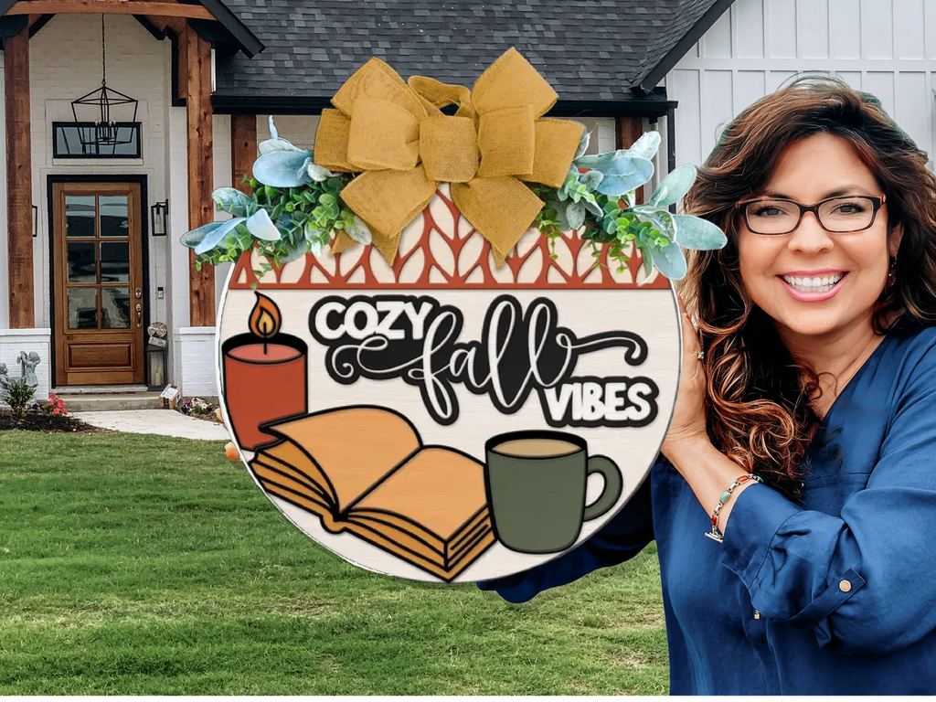 A smiling woman with wavy brown hair and glasses holds a decorative sign reading Cozy Fall Vibes with images of a candle, book, and mug in front of a house with a green lawn.