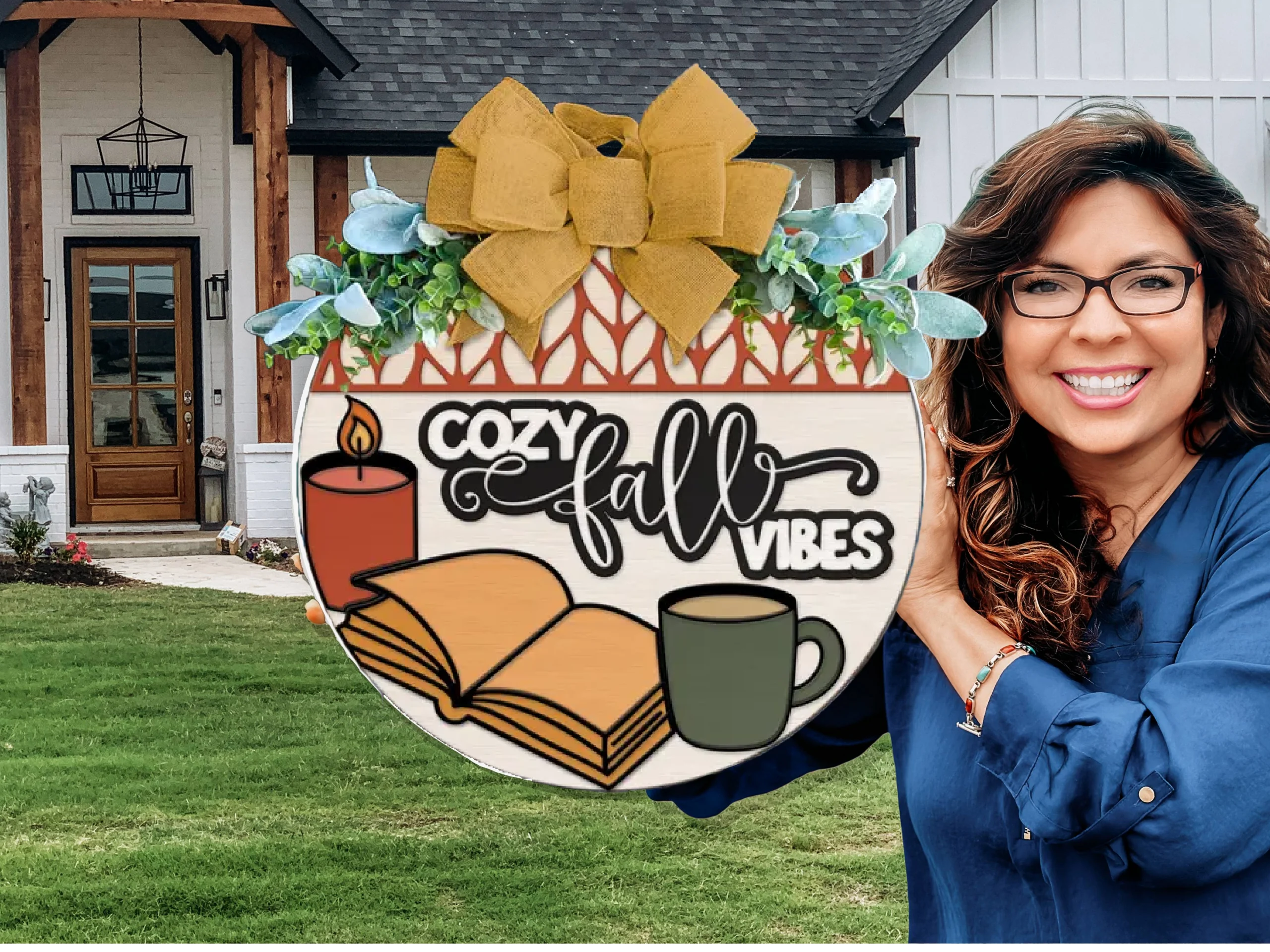 A smiling woman with wavy brown hair and glasses holds a decorative sign reading Cozy Fall Vibes with images of a candle, book, and mug in front of a house with a green lawn.