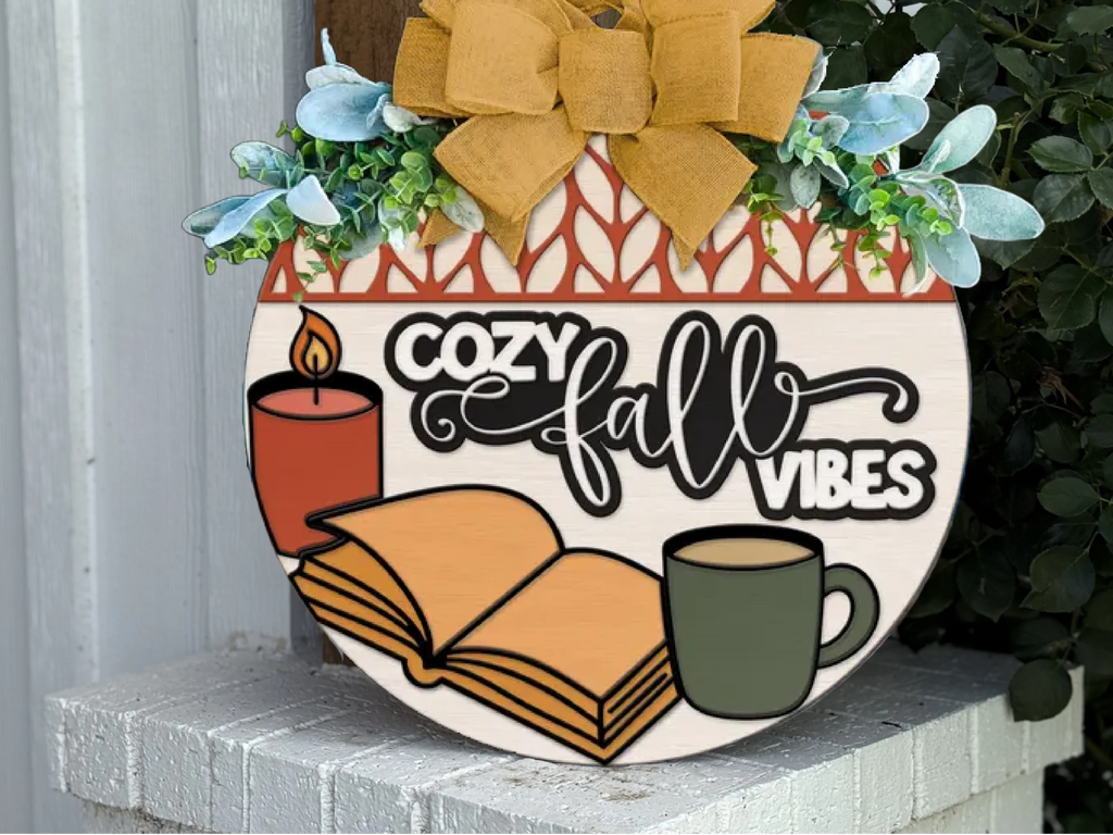 A round sign decorated with greenery and a yellow bow reads Cozy Fall Vibes and features images of a lit candle, an open book, and a green mug. The sign sits on a white brick surface near plants.