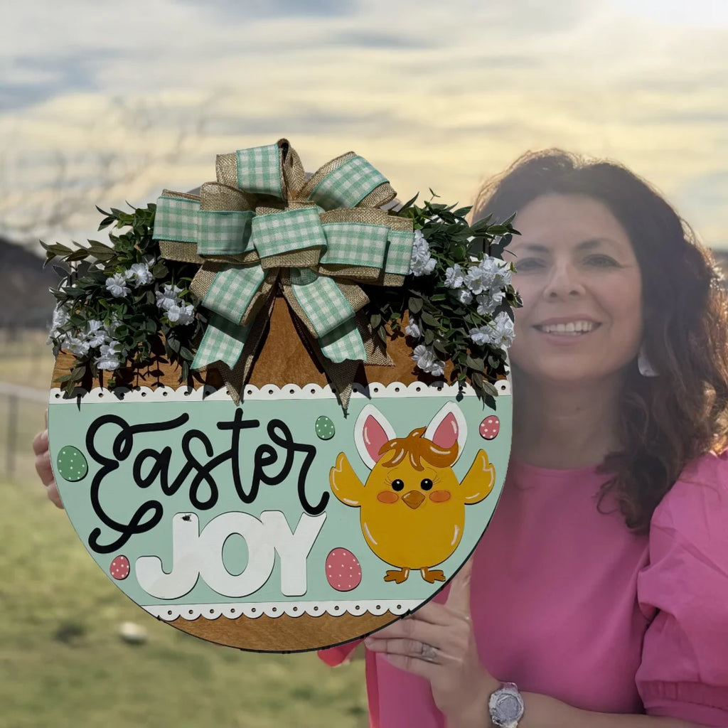 A woman in a pink blouse holds a round Easter wreath. The wreath features greenery, white flowers, a plaid bow, and a decorative sign with Easter Joy written on it. A cartoon yellow chick with bunny ears is depicted next to colorful Easter eggs. The background shows a field and cloudy sky.