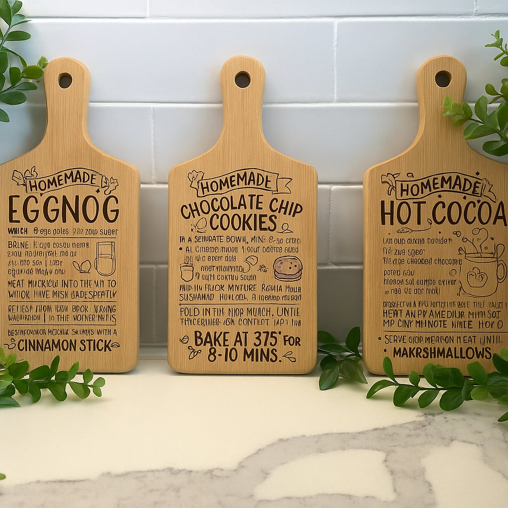 Three Engraved Recipe Cutting Boards (11” or 15.5”) featuring handwritten recipes for eggnog, apple pie, and hot cocoa stand on a kitchen counter, accented by green plants and a white tile backsplash.