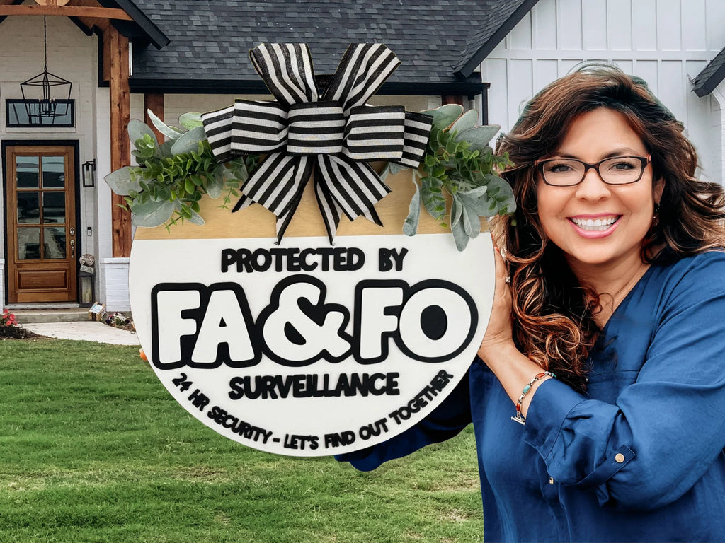 A woman with long, wavy hair and glasses smiles while holding a circular sign. The sign displays bold text: Protected by FA & FO Surveillance, and adds, 24 Hr Security - Lets Find Out Together. The scene is outdoors, in front of a house with a lawn.