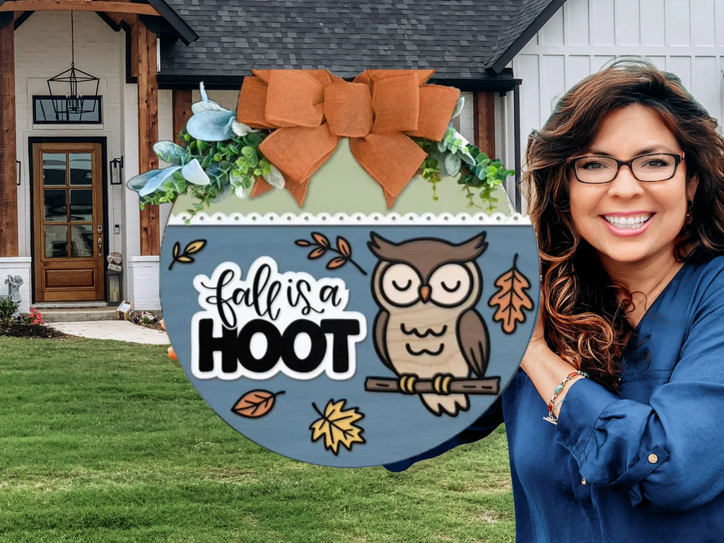 A smiling woman stands in front of a house holding a round fall-themed sign decorated with an owl, leaves, and the words fall is a HOOT, topped with a large orange bow and greenery.