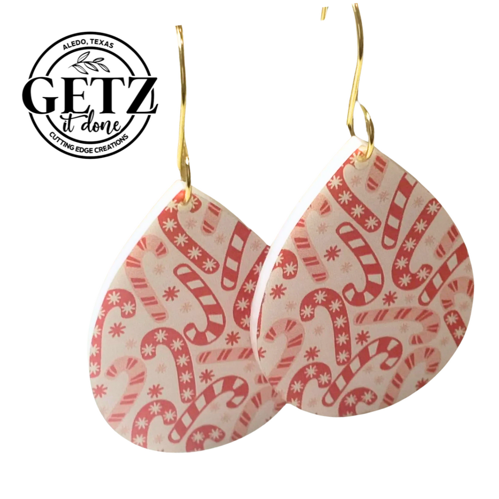 Teardrop-shaped earrings with a red and white candy cane pattern, featuring small snowflakes. The earrings have gold hooks. The background is white, and the logo in the corner reads GETZ it done, Aledo, Texas, Cutting Edge Creations.