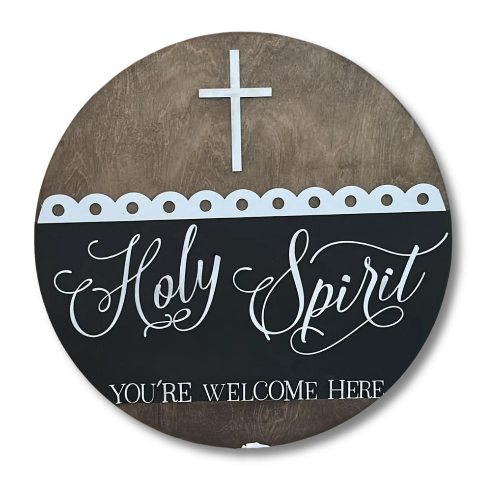 A round wall decoration with a wooden texture background features a white cross at the top. Below, it says Holy Spirit in elegant script and YOURE WELCOME HERE in block letters, divided by a scalloped white border.