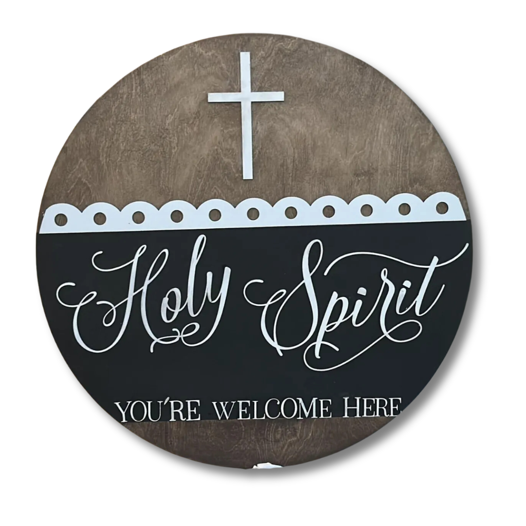 A round wall decoration with a wooden texture background features a white cross at the top. Below, it says Holy Spirit in elegant script and YOURE WELCOME HERE in block letters, divided by a scalloped white border.