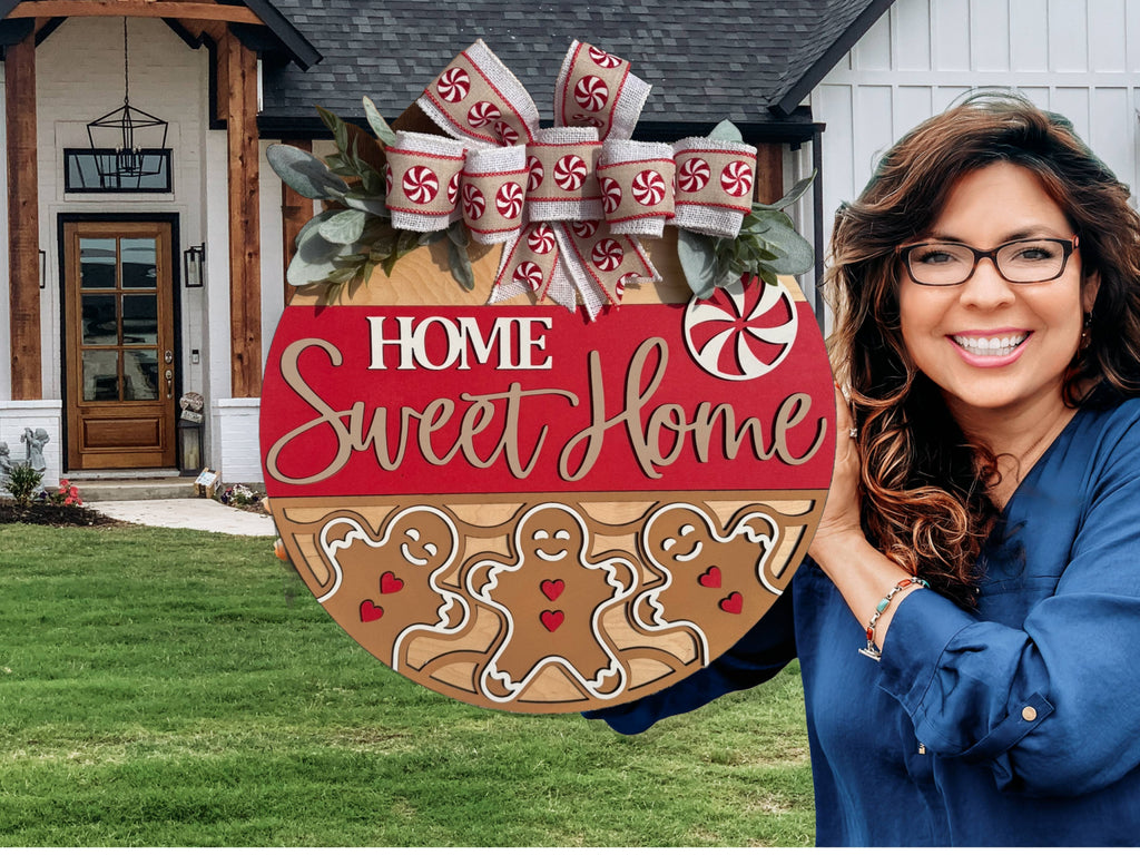 A smiling woman in glasses stands outside a modern house, holding the Welcome To The Nuthouse Nutcracker Sign Welcome Door Sign, decorated with festive nutcracker designs and a bow.