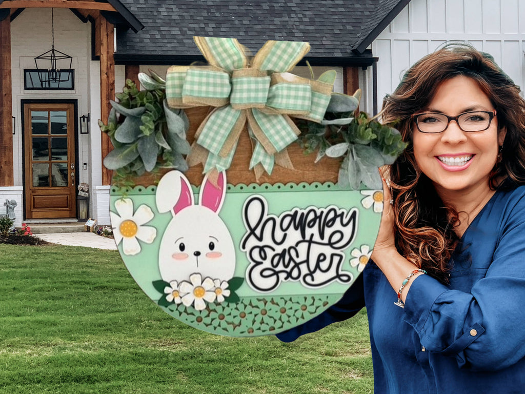 A smiling woman with wavy brown hair and glasses holds a round, decorative Easter sign with a bunny, flowers, greenery, and a bow. The sign reads happy Easter and is displayed in front of a modern house.