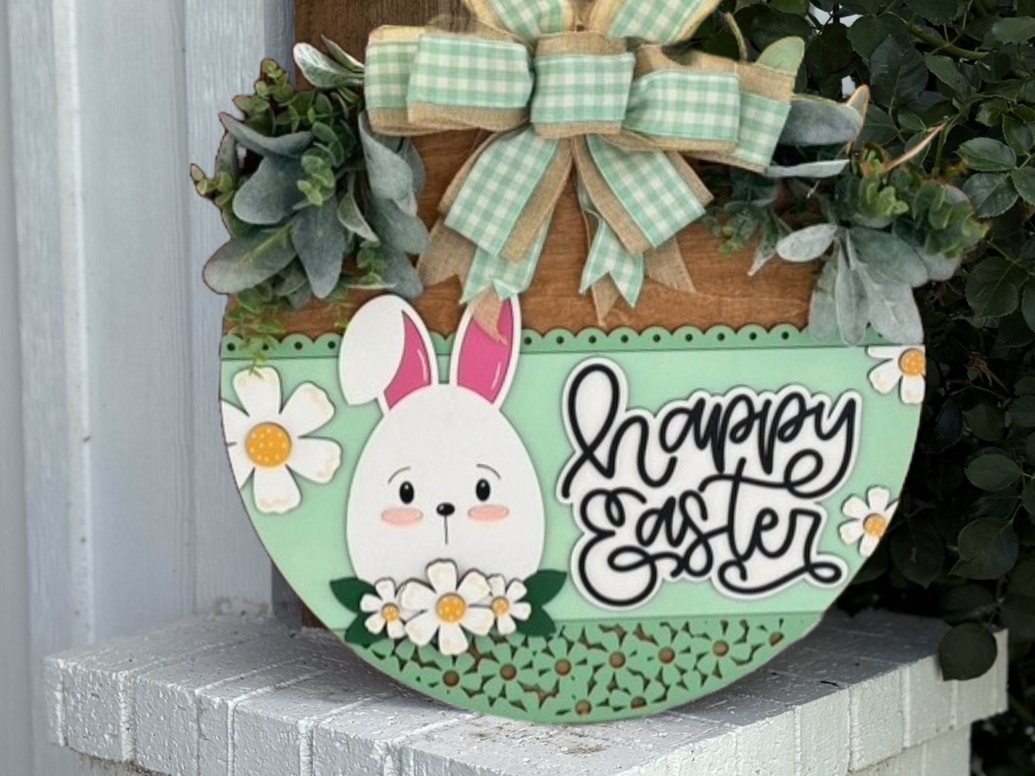 A round wooden Easter sign decorated with a cartoon bunny, white daisies, green leaves, and a green plaid bow. The sign reads happy Easter in cursive lettering and is propped on a white brick surface.