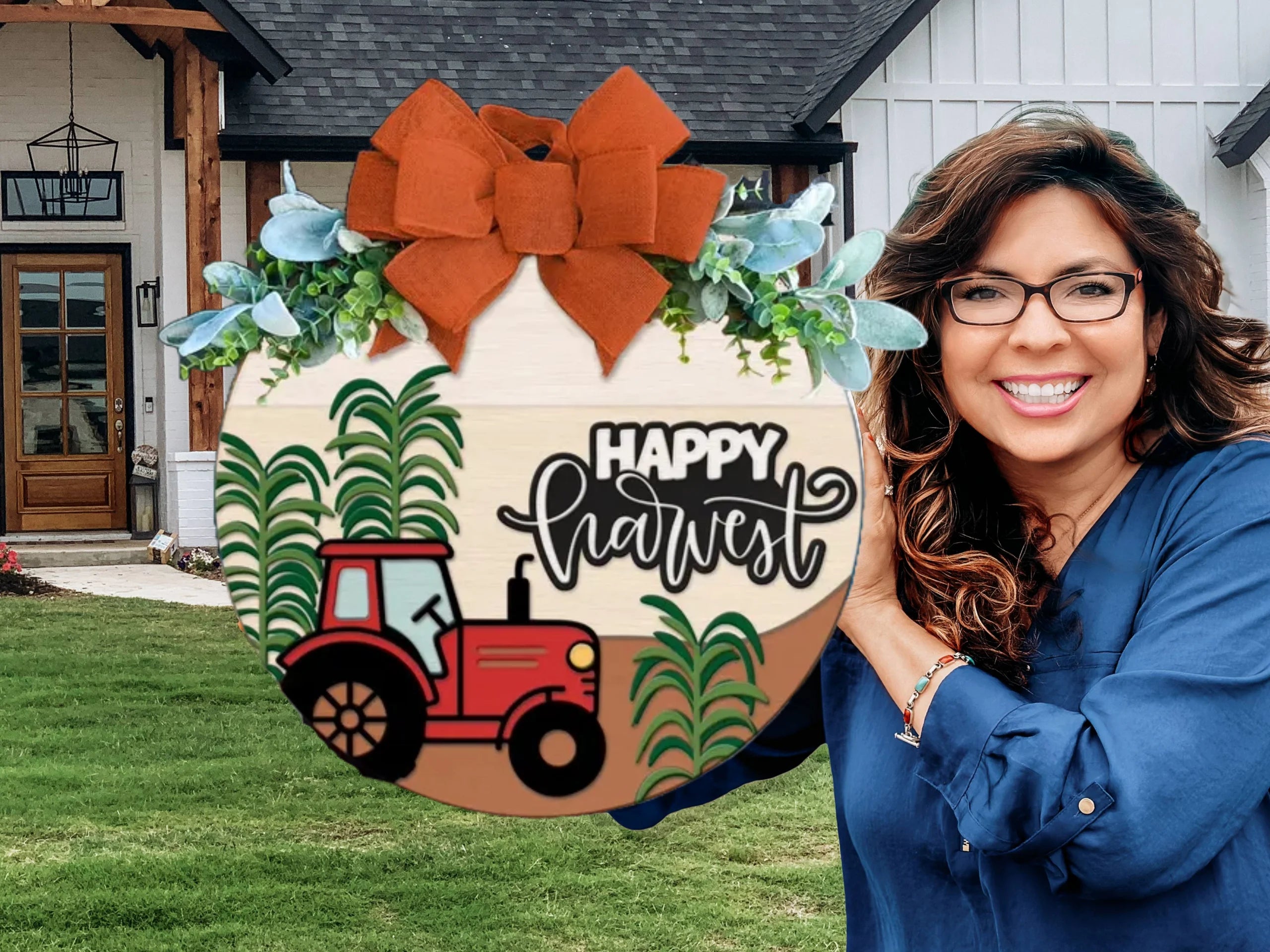 A smiling woman in glasses holds a round “Happy Harvest” sign with a red tractor, green plants, and a large orange bow in front of a modern farmhouse.
