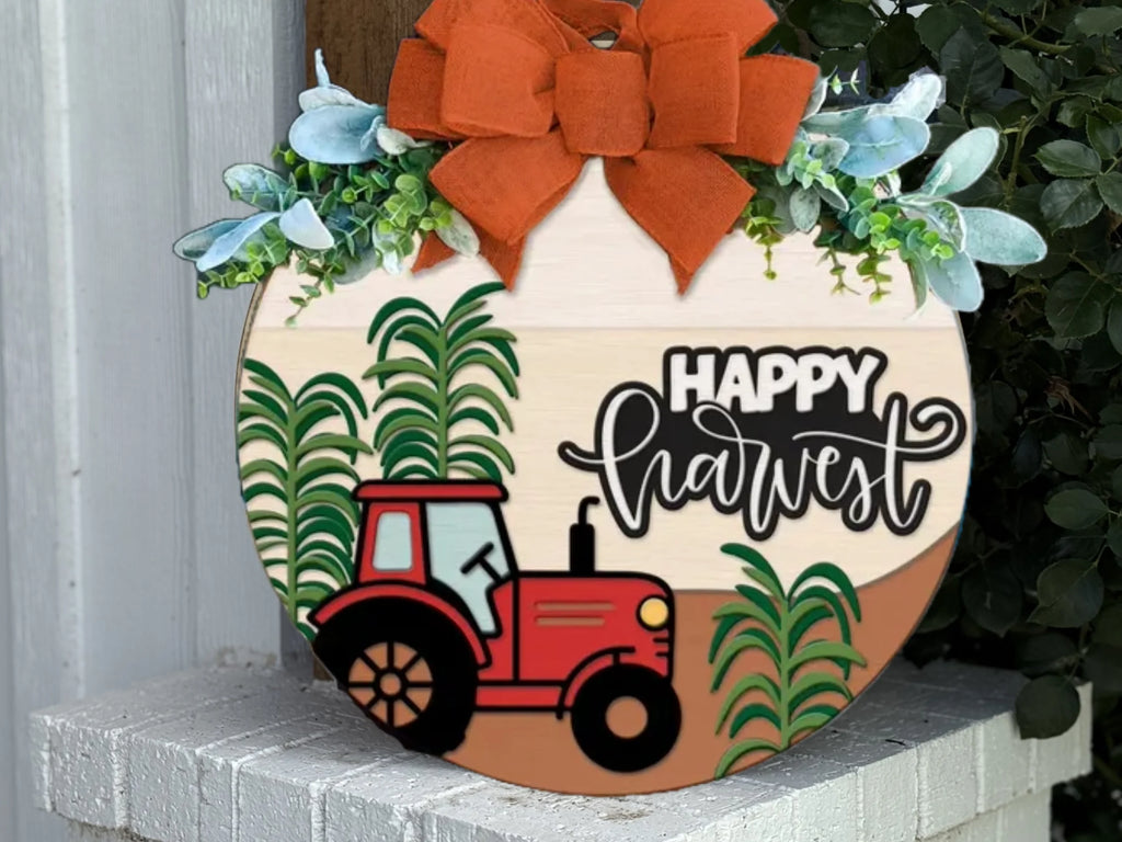 A round wooden sign decorated with an orange bow and greenery, featuring a red tractor, green crops, and the words “Happy Harvest” in black and white script. The sign sits on a white brick ledge near some plants.