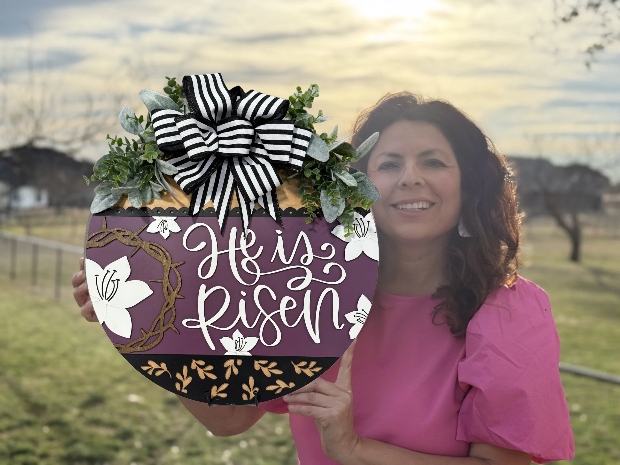 Woman in a pink top holds a round Easter decoration with the phrase He is Risen in white script. The sign features a large black and white striped bow, greenery, and floral accents. The background shows a sunlit field with bare trees, suggesting early spring.