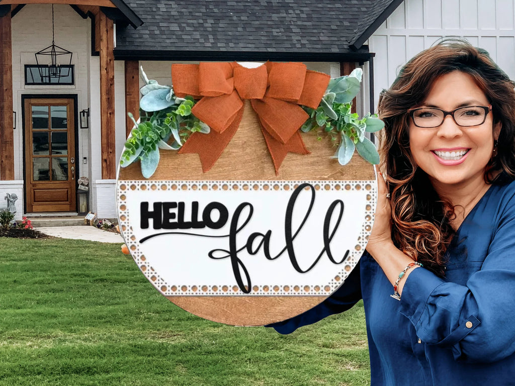 A smiling woman with long brown hair and glasses holds a decorative round wooden sign that says Hello Fall with greenery and a large orange bow, standing in front of a modern house with a lawn.