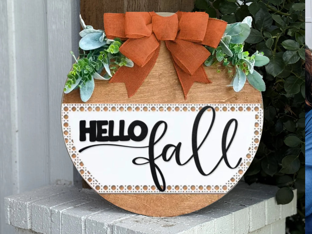A round wooden sign with greenery and an orange bow on top, reading HELLO fall in black script, is displayed on a white brick surface near some green foliage.