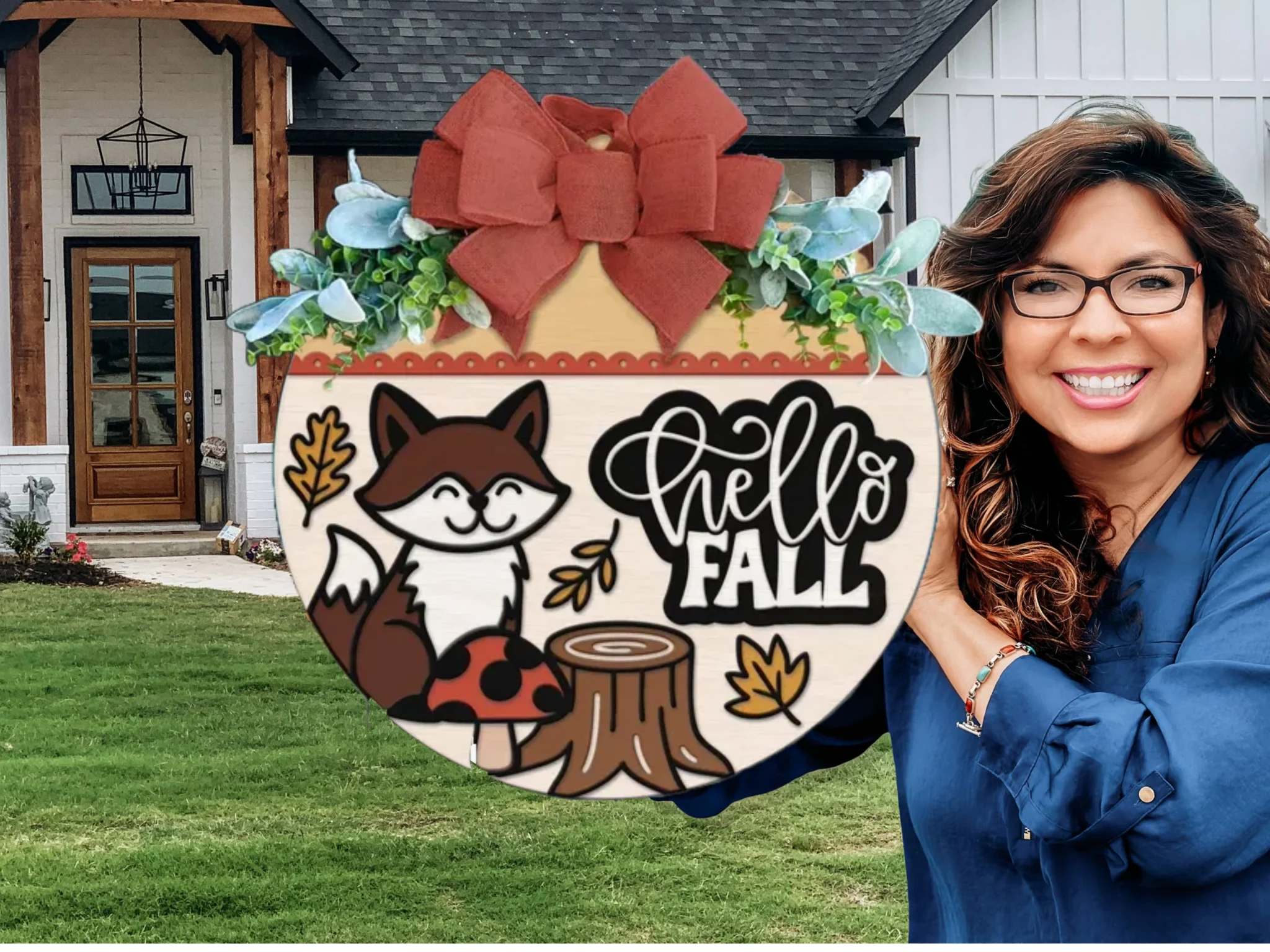 A smiling woman with glasses holds a round fall-themed sign featuring a fox, stump, leaves, and the words Hello Fall, decorated with greenery and a red bow, standing outside a modern house.