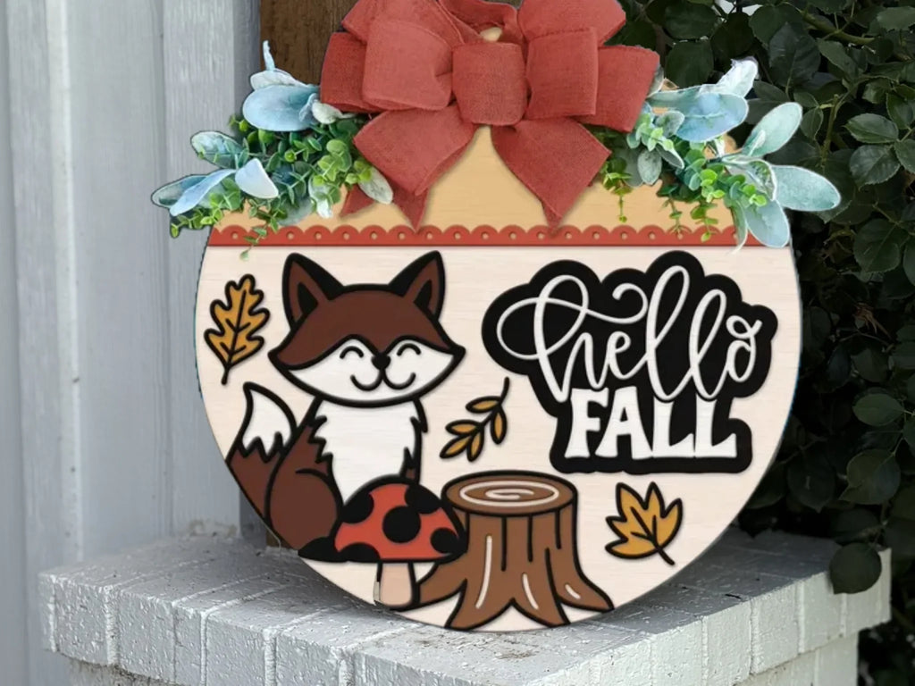 A round wooden sign with a red bow, leaves, and flowers on top, featuring a smiling fox, a tree stump, leaves, a mushroom, and the words “hello FALL” in bold script, displayed on a white brick surface.