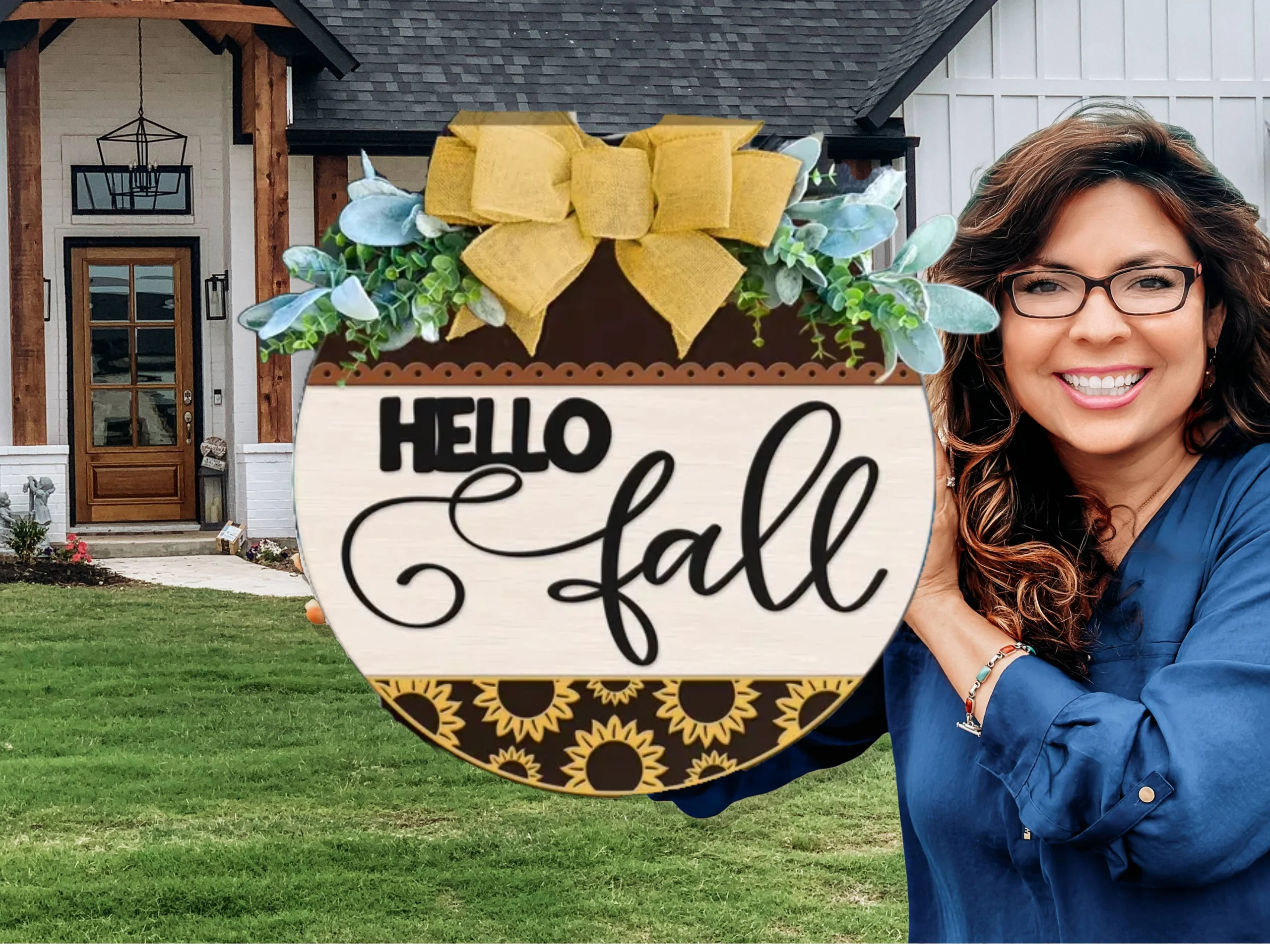 A smiling woman in glasses holds a decorative round sign with flowers, a yellow bow, and the words Hello fall in front of a house with a green lawn.