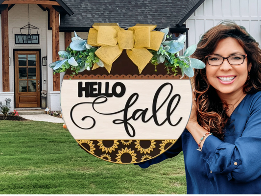 A smiling woman in glasses holds a decorative round sign with flowers, a yellow bow, and the words Hello fall in front of a house with a green lawn.