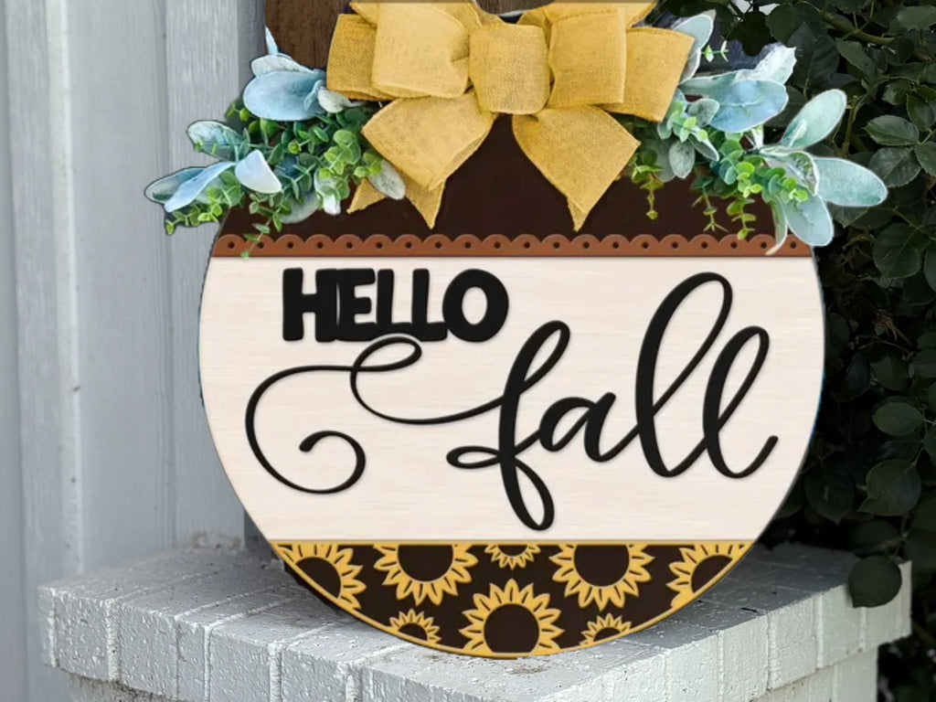 A round wooden sign with HELLO fall written on it, decorated with sunflowers at the bottom, greenery and a large yellow bow at the top, leaning against a white brick wall.
