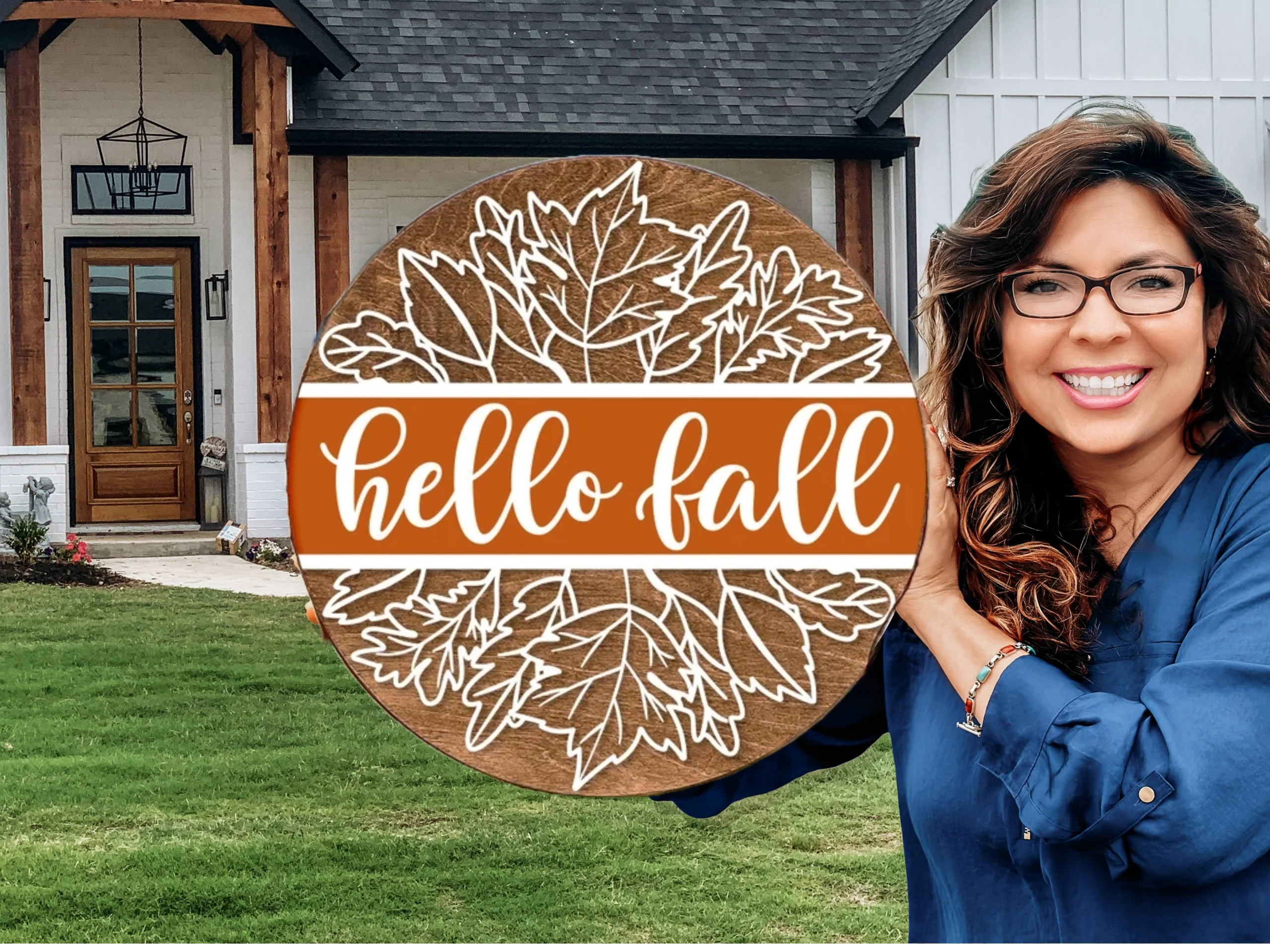 A smiling woman in glasses and a blue shirt holds a round wooden sign with white outlined leaves and the words hello fall in front of a modern house with a lawn.
