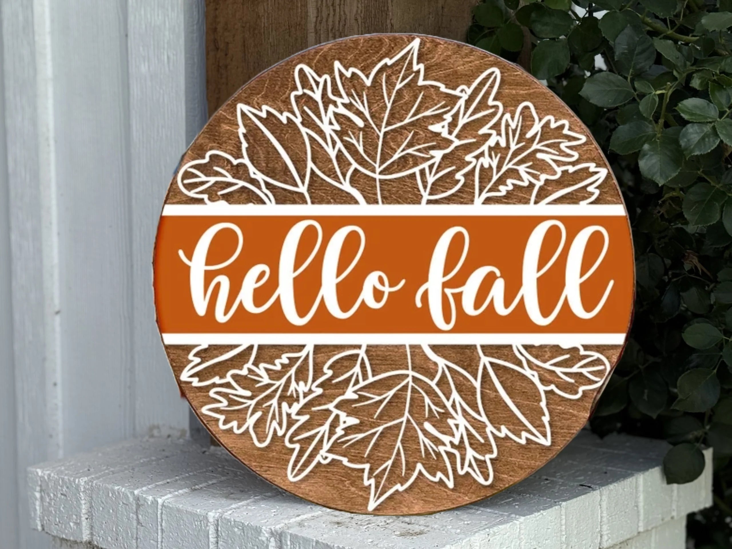 A round wooden sign with white outlined autumn leaves and an orange band in the center that reads hello fall in cursive white letters. The sign is displayed outside near greenery.