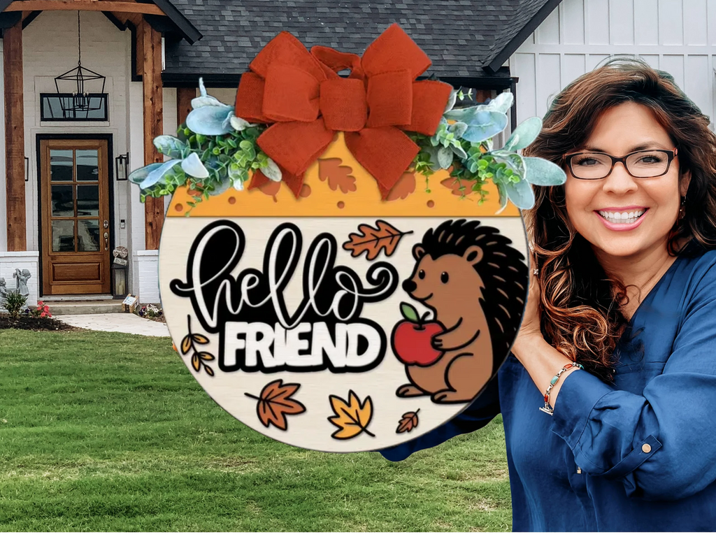 A smiling woman holds a round autumn-themed sign that says hello FRIEND with a cartoon hedgehog, leaves, and an apple. She stands in front of a modern house with white siding and wood accents.