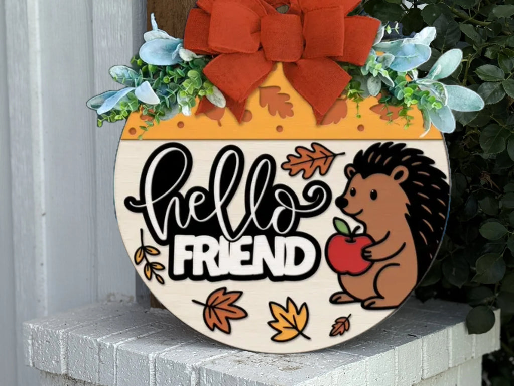 A round decorative sign with a red bow, green leaves, and autumn accents. It says hello FRIEND and shows a smiling hedgehog holding an apple, surrounded by fall leaves.