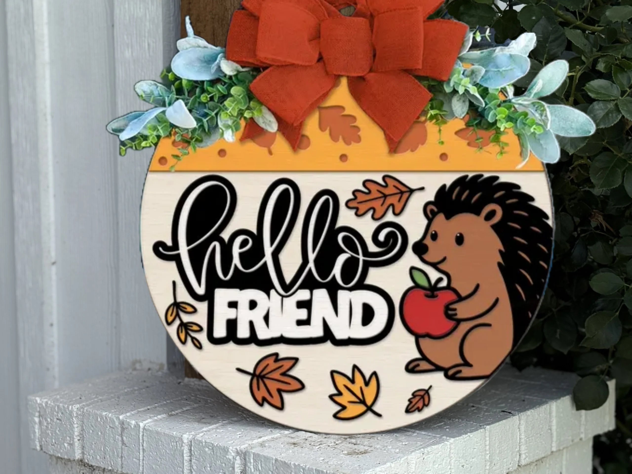 A round decorative sign with a red bow, green leaves, and autumn accents. It says hello FRIEND and shows a smiling hedgehog holding an apple, surrounded by fall leaves.