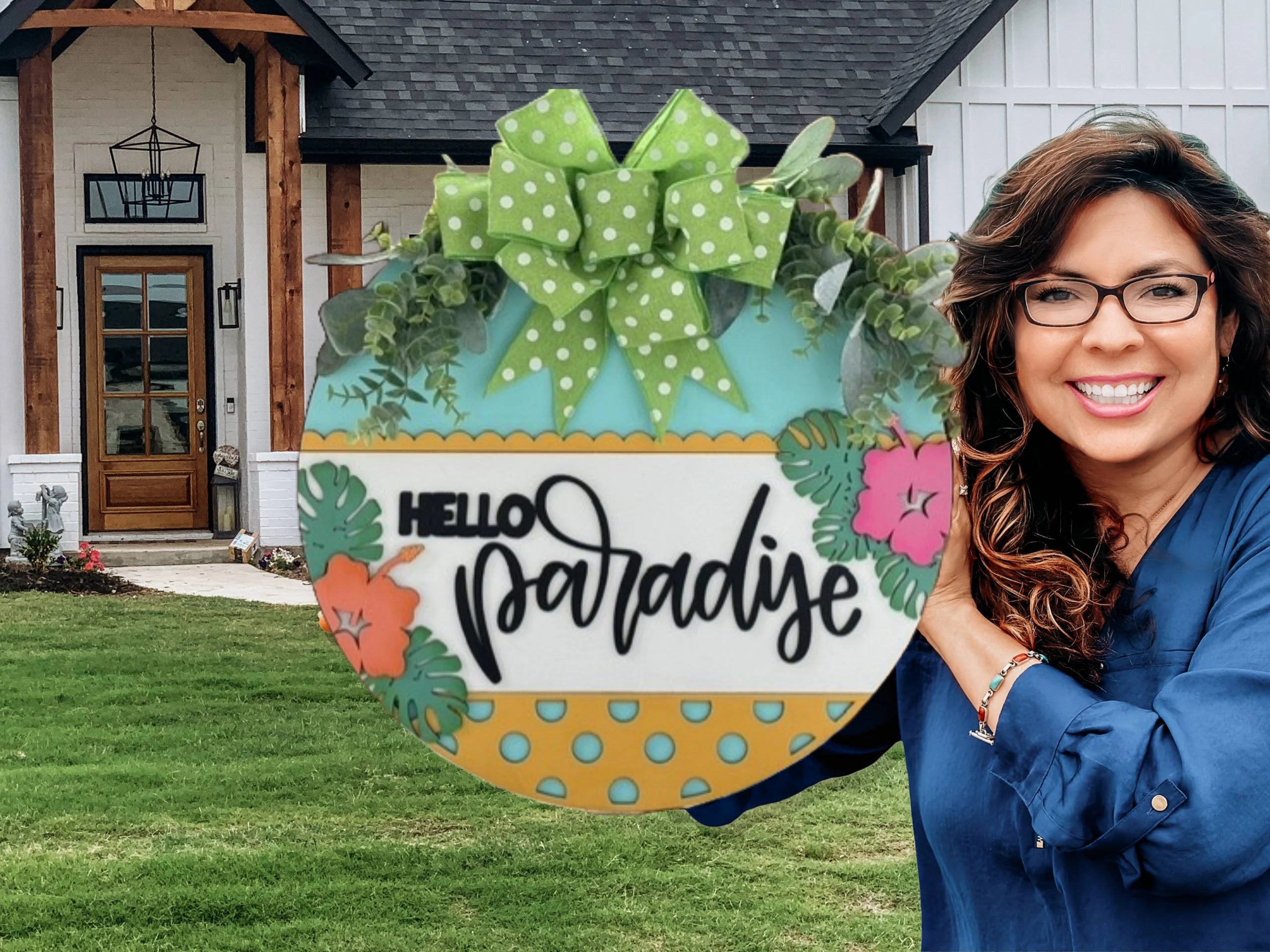 Smiling woman with long wavy brown hair and glasses holds a decorative circular sign reading Hello Paradise, featuring a green bow and tropical designs. Shes standing in front of a modern house with a wooden front door and green lawn.