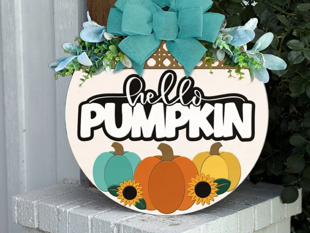 Round autumn-themed sign with a teal bow, greenery, and the words hello PUMPKIN. The sign features three pumpkins in teal, orange, and yellow, accented by two sunflowers at the bottom.