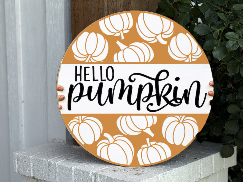A round sign decorated with orange and white pumpkins and the words HELLO pumpkin in black script is held by two small hands, one on each side. The sign rests on a white brick ledge near some green leaves.