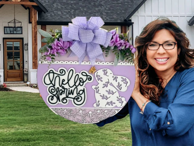 A smiling woman in a blue shirt holds a circular lavender sign that reads Hello Spring in script. The sign is decorated with purple flowers, a bow at the top, and a white floral pattern. She stands in front of a house with a grass lawn and wooden front porch accents.