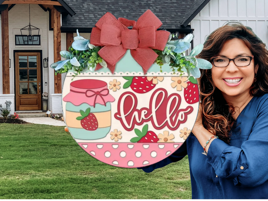 A smiling woman with long brown hair and glasses holds a large, round sign decorated with strawberries, jam jars, a red bow, and the word hello in front of a modern house with a lawn.