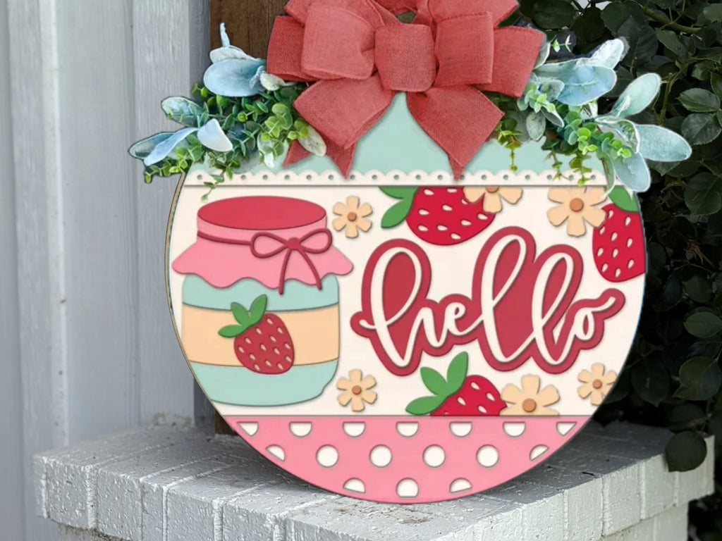 A round decorative sign with strawberries, a jam jar, flowers, polka dots, and the word hello in red script, topped with a red bow and greenery, hanging on a white brick wall.