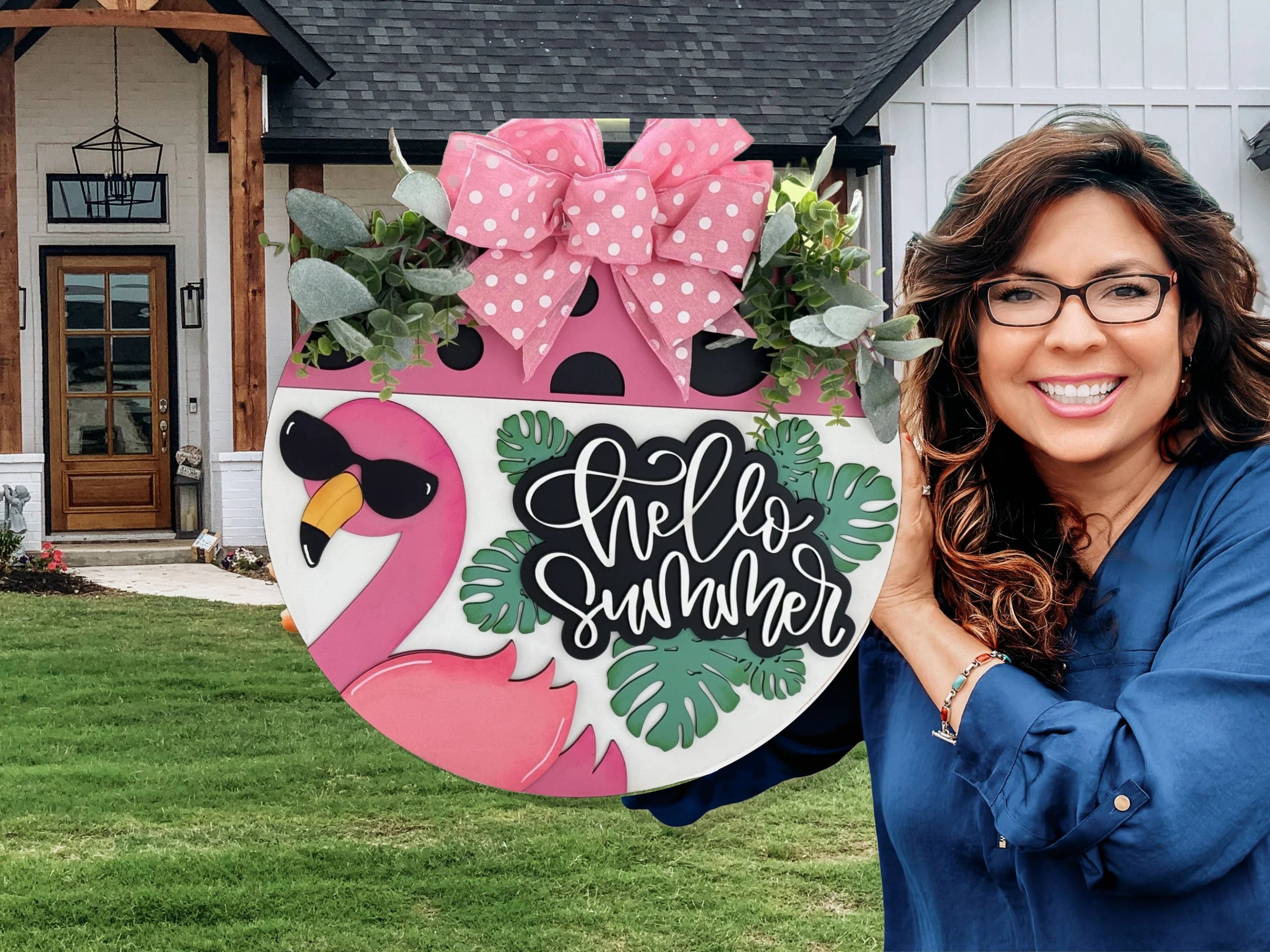 Smiling woman with long brown hair and glasses, wearing a blue blouse. She holds a round sign featuring a pink flamingo, tropical leaves, and the text Hello Summer in cursive. The sign is topped with a large pink bow. In the background is a house with a green lawn.