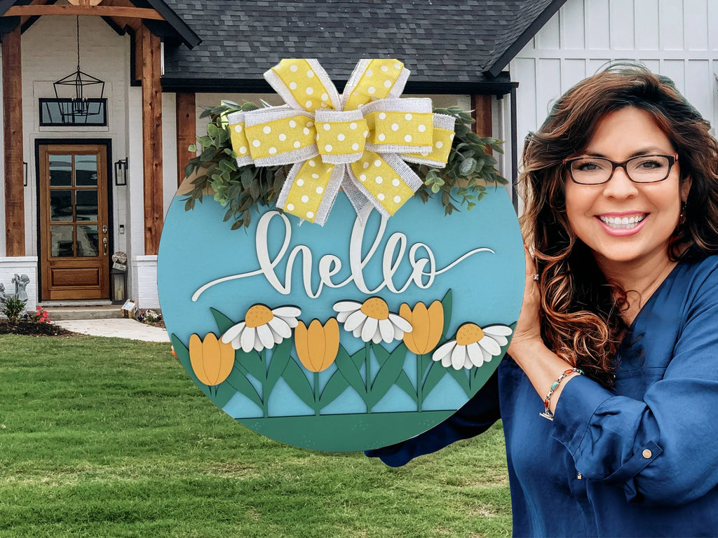 Smiling woman in a blue shirt holds a colorful circular sign with yellow tulips, white daisies, and the word hello in white script. A large yellow and white polka dot bow tops the sign. A modern house with wooden accents and a manicured lawn is in the background.