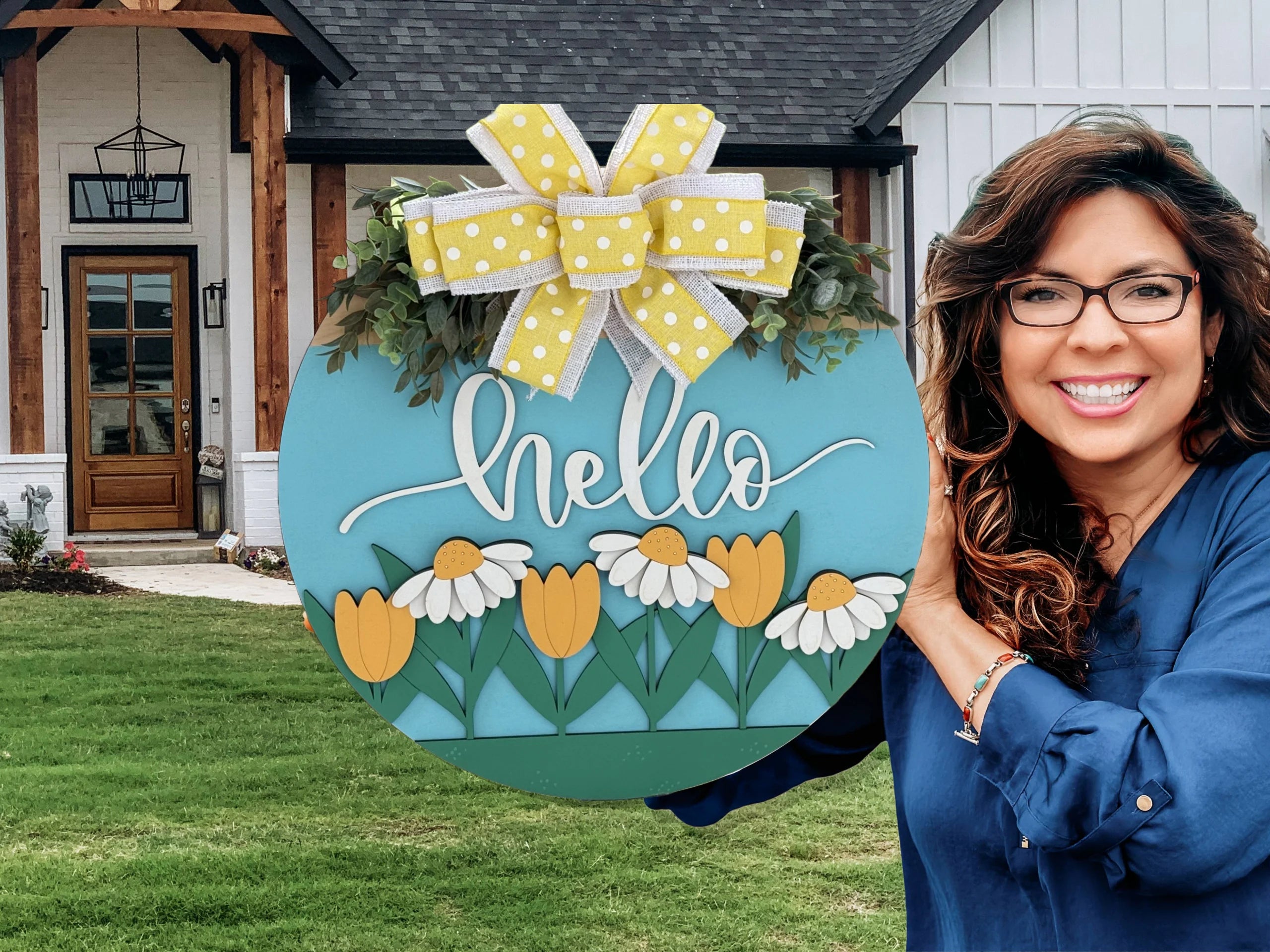 Smiling woman in a blue shirt holds a colorful circular sign with yellow tulips, white daisies, and the word hello in white script. A large yellow and white polka dot bow tops the sign. A modern house with wooden accents and a manicured lawn is in the background.
