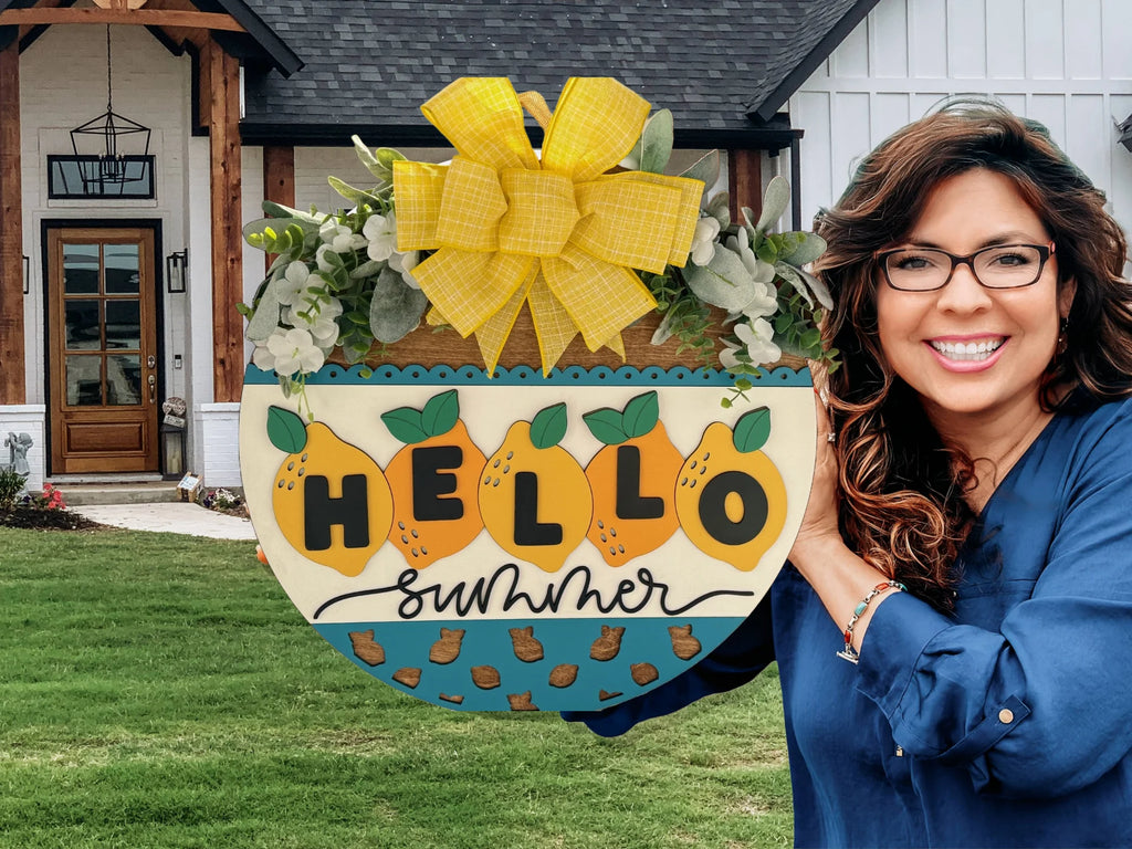 Woman smiling and holding a decorative sign that reads Hello Summer with lemon motifs and a large yellow bow at the top. She stands in front of a modern house with a gabled roof and wooden porch. The lawn is green and well-maintained.