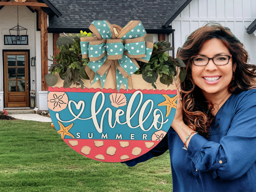 A smiling woman in a blue shirt is holding a round, decorative sign. The sign is painted with hello SUMMER in white and features stars, hearts, and polka dots. It has a large bow and greenery at the top. In the background is a white house with a wooden door and grassy lawn.