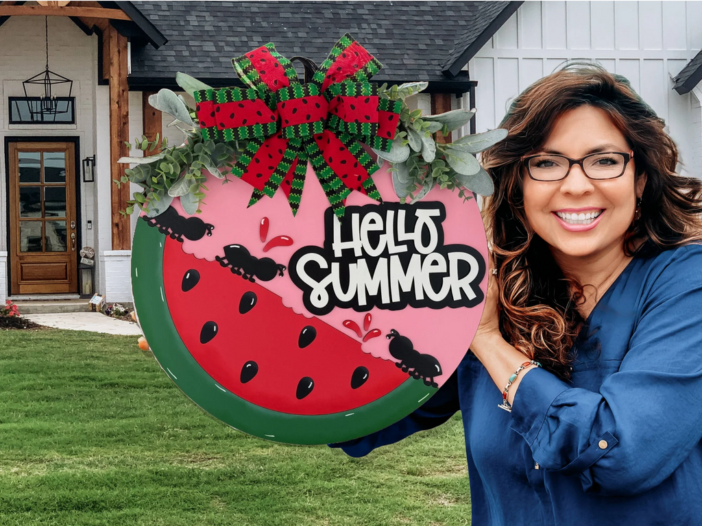 Smiling woman in a blue shirt holds a decorative sign shaped like a watermelon slice, featuring a red and green bow, faux leaves, and ants. The sign reads Hello Summer in bold white letters. A house with a black roof and wooden accents is in the background.