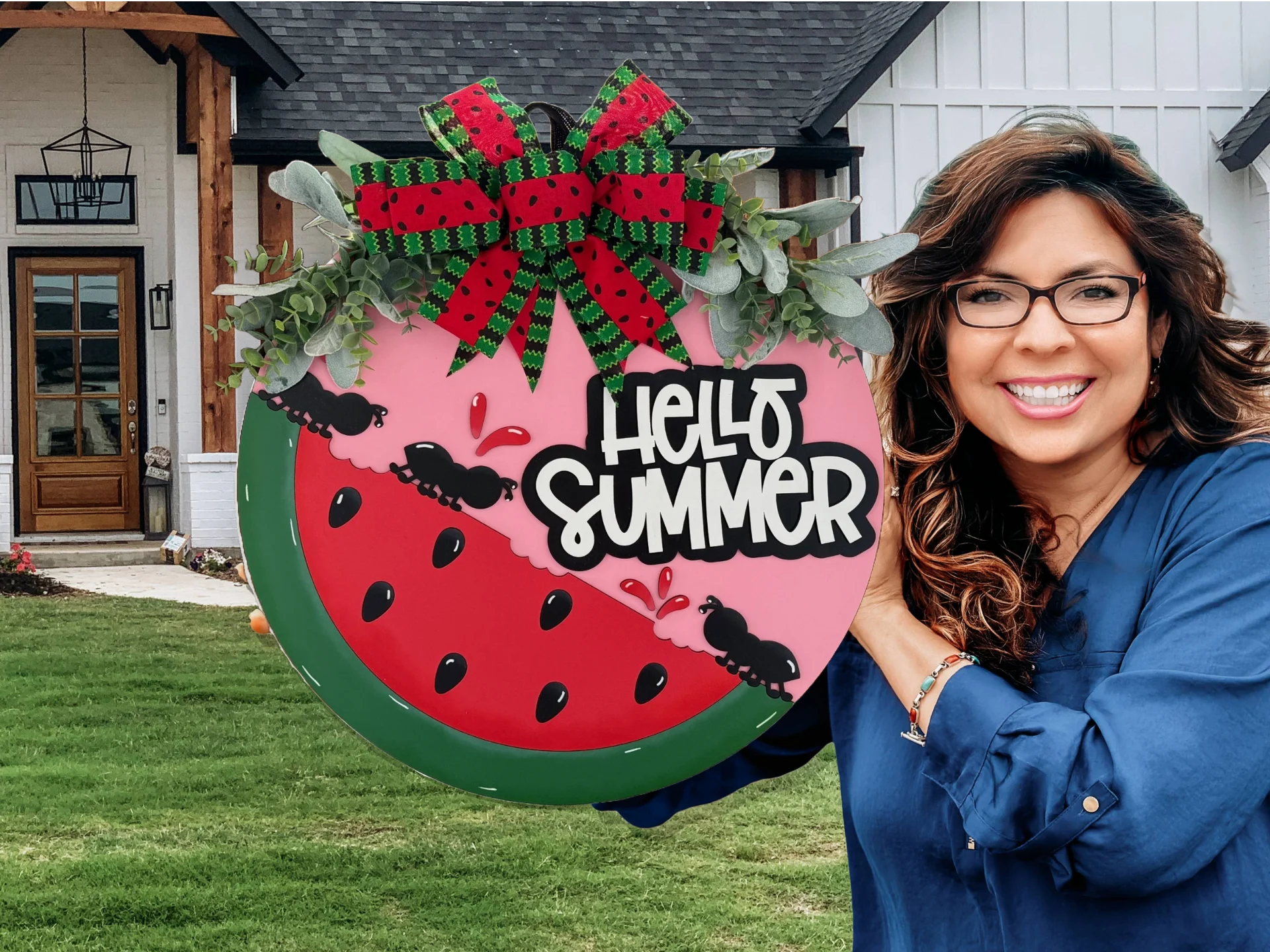Smiling woman in a blue shirt holds a decorative sign shaped like a watermelon slice, featuring a red and green bow, faux leaves, and ants. The sign reads Hello Summer in bold white letters. A house with a black roof and wooden accents is in the background.