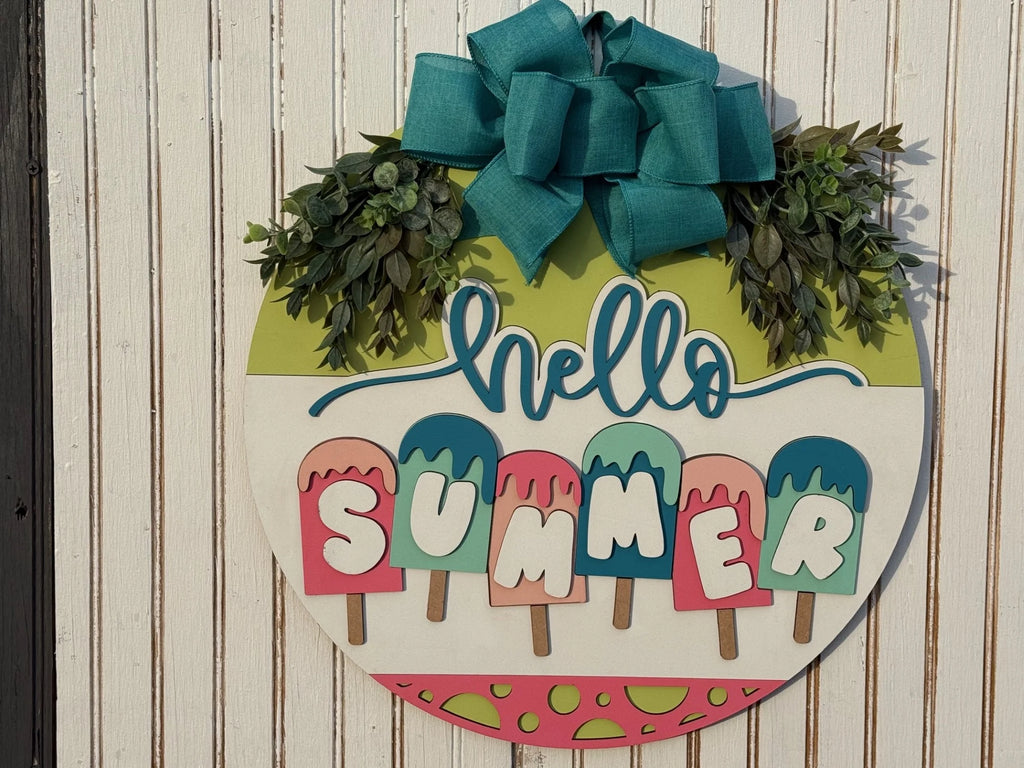 The Home Sweet Home Door Sign features colorful letters spelling hello SUMMER like popsicles, with a teal bow and green foliage above, creating a vibrant and lively summery theme in green and pink hues on a white wall.