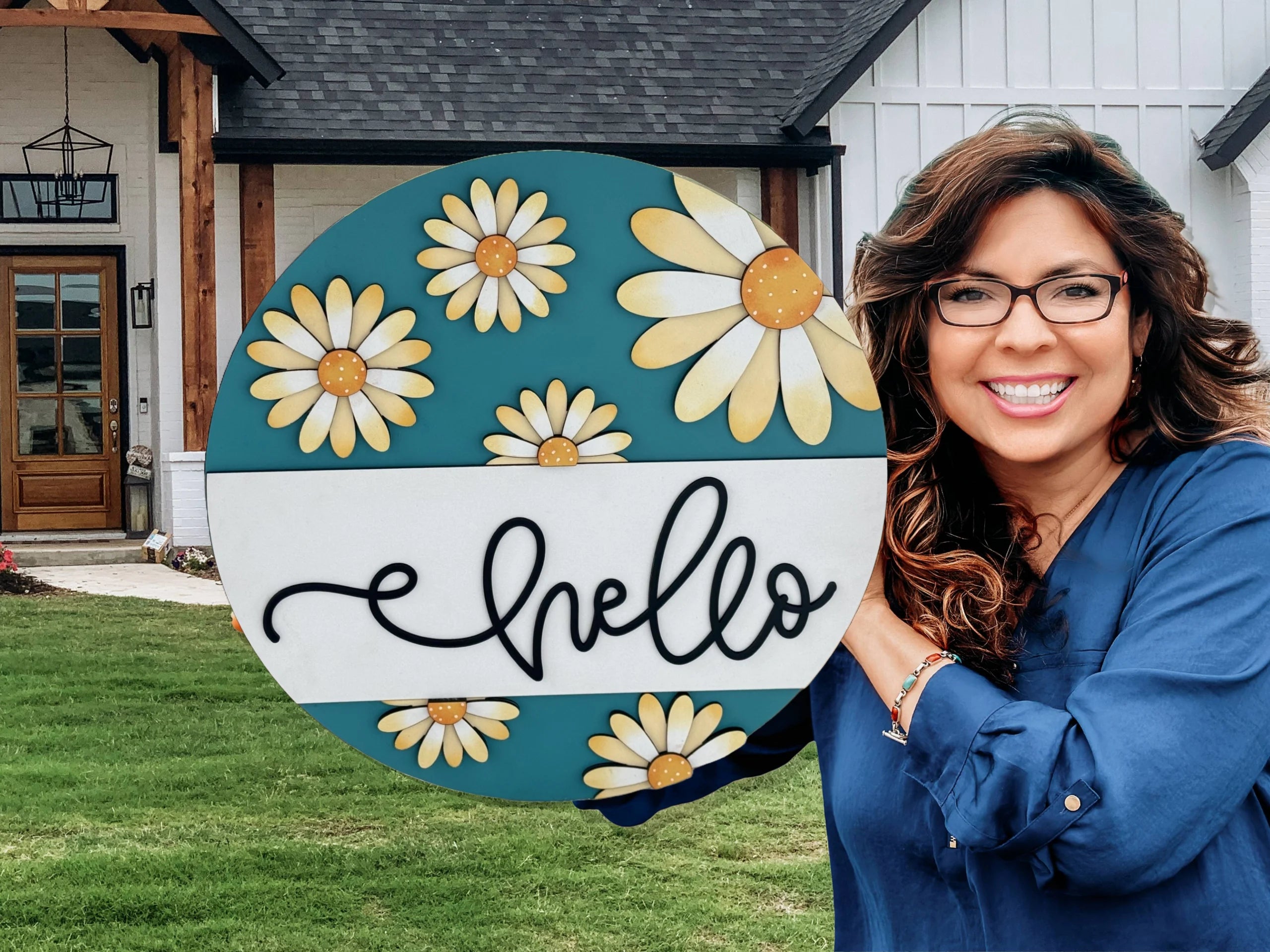 Woman in a blue blouse smiles while holding a round decorative sign featuring yellow and white daisies and the word hello in cursive. She stands in front of a modern house with a grassy lawn and wooden front door.