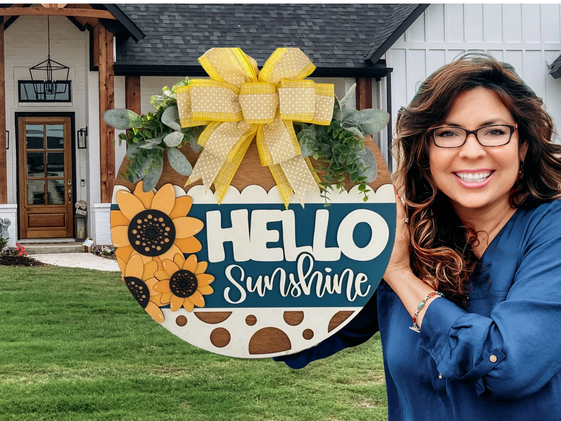 A woman in a blue shirt smiles while holding a round, decorative sign that says Hello Sunshine with sunflower designs and a yellow polka dot ribbon. In the background, theres a house with white siding and a wooden door. The lawn is green and well-maintained.