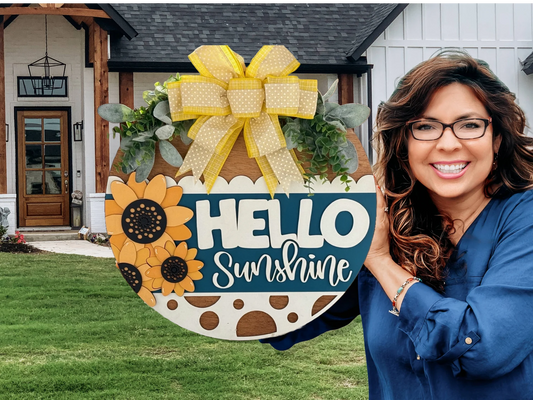 A woman in a blue shirt smiles while holding a round, decorative sign that says Hello Sunshine with sunflower designs and a yellow polka dot ribbon. In the background, theres a house with white siding and a wooden door. The lawn is green and well-maintained.