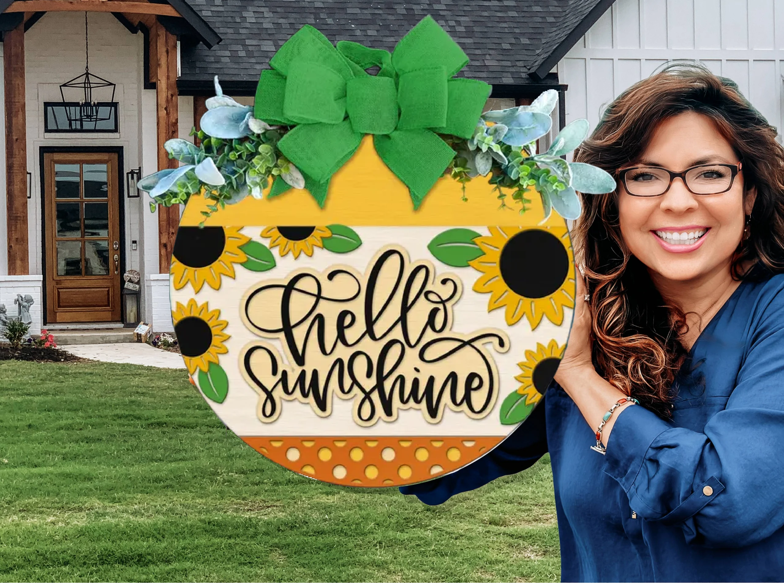 A woman with wavy brown hair and glasses smiles while holding a round wooden sign decorated with sunflowers, greenery, a green bow, and the words hello sunshine in front of a modern house with a lawn.
