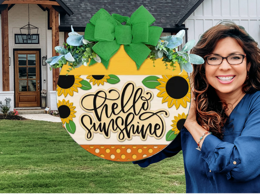 A woman with wavy brown hair and glasses smiles while holding a round wooden sign decorated with sunflowers, greenery, a green bow, and the words hello sunshine in front of a modern house with a lawn.
