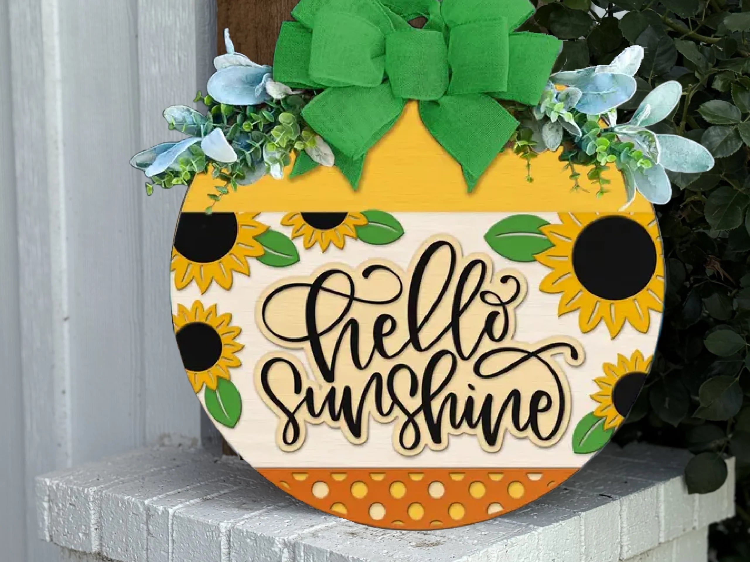 A round wooden sign decorated with sunflowers, green leaves, and a large green bow displays the words hello sunshine in cursive. The sign rests on a white brick surface near a white wall.