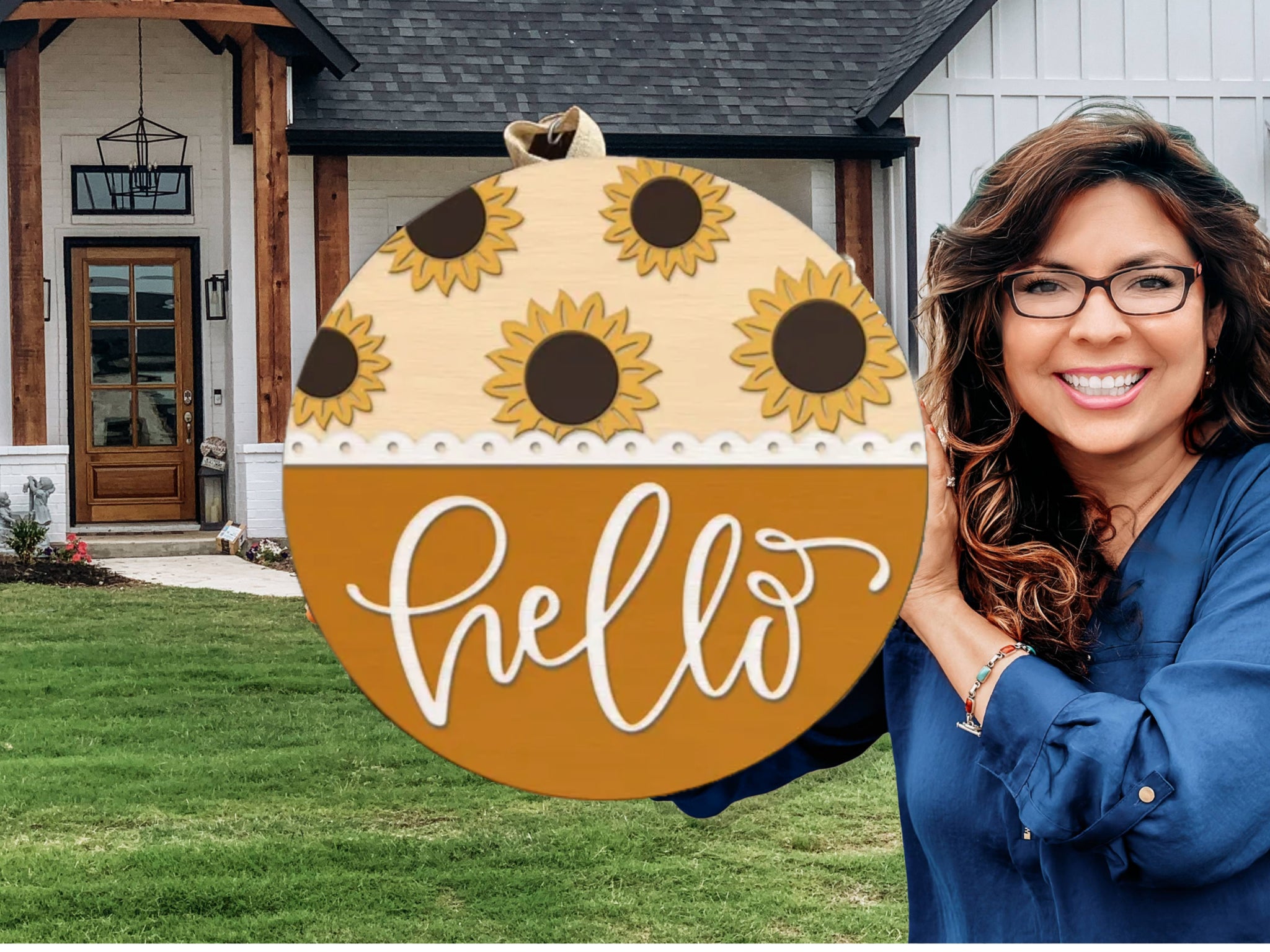 A smiling woman with long brown hair and glasses stands outside a house, holding a round wooden sign decorated with sunflowers and the word “hello” written in white script.